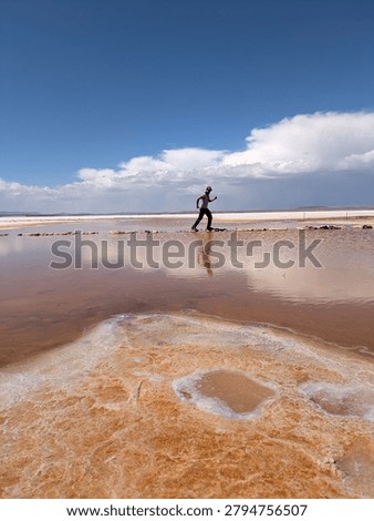 Man balancing on stone path across water pool at Salar de Uyuni salt flat in Bolivia, tourist exploring pink mineral water and white desert landscape under blue sky, South America travel