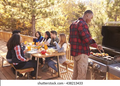 Man barbecues for friends at a table, on a deck in a forest