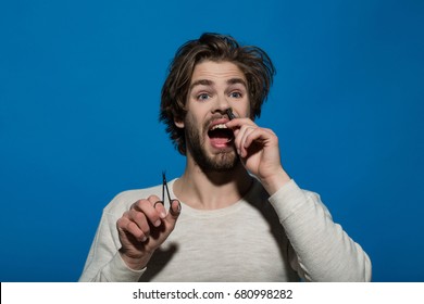 Man Barber With Surprised And Scared Face Twitch Nose Hair With Tweezers And Hold Scissors, Guy Has Long Hair And Beard On Blue Background