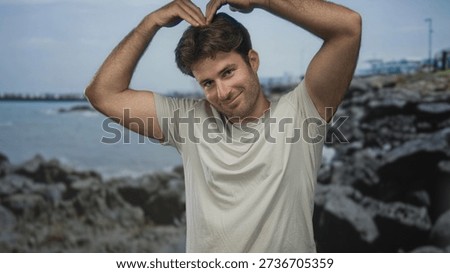 Man with bare arms forms heart with hands above head on rocky seaside street, smiling at camera; affection warmth.