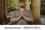 Man with bare hands pressed together in prayer under stone columns on an old town street looking upward; quiet faith.