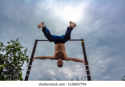 A man with a bare torso in blue pants upside down doing his morning exercises on a horizontal bar - Shutterstock ID 1136037794