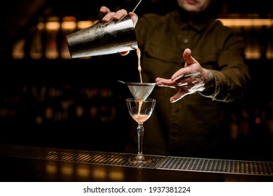 man bartender carefully filters bright alcoholic cocktail from steel shaker cup into glass through sieve. Close-up view.