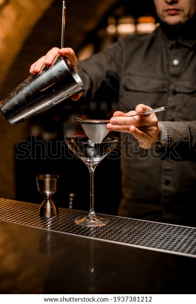 man bartender holds in his\
hands sieve and steel shaker cup over empty transparent martini\
glass