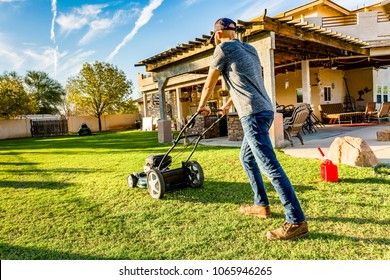 Man in baseball cap mowing green lawn on bright summer day in backyard. Red gasoline can sits nearby on grass ready to refuel mower. 