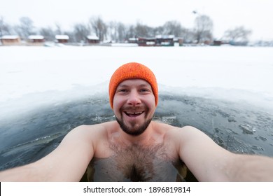 A man bathes in a river ice hole in winter. Orange hat. Selfie. Smile. Day, cloudy.