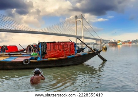 Man bathing in the Ganges river with view of wooden boats and Vidyasagar setu bridge at sunrise at Kolkata, India