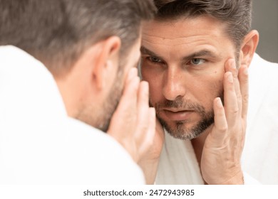 a man in a bathrobe in the bathroom looking at his wrinkles and pimples on his face. A man is looking at his reflection in a mirror. A man is looking at his reflection in a mirror.  - Powered by Shutterstock