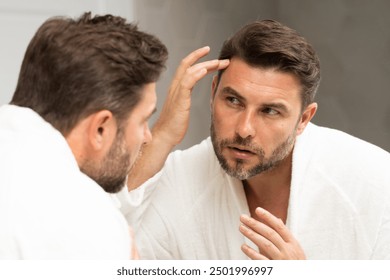 a man in a bathrobe in the bathroom looking at his wrinkles and pimples on his face. A man is looking at his reflection in a mirror. A man is looking at his reflection in a mirror.  - Powered by Shutterstock