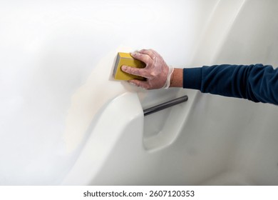 Man in bathroom tub wall white painted plastic doing remodeling renovation cleaning, resurfacing sanding the peeling paint