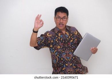 A man in batik shirt holds a laptop while shows an ok sign with his fingers; excited, high-spirited expression.