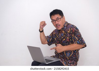 A man in batik shirt pointing at his laptop, clenching his fist; excited, expressing things were done successfully.
