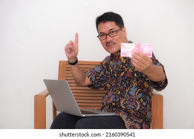 A man in batik shirt shows one hundred thousand rupiah bill, a computer on his lap; smiling. 