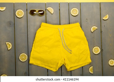 Man Beach Accessories On Wooden Background. Yellow Swim Trunks, Aviator Sunglasses Between Parts Of Lemons On Grey Wooden Board.