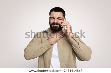 Man with a beard appears distressed while talking on the phone, hand on throat suggesting difficulty speaking or a sore throat, in a beige shirt against a plain background