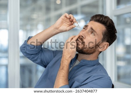 A man with a beard applying eye drops to his eye, likely suffering from eye irritation. He appears to be in pain or discomfort.