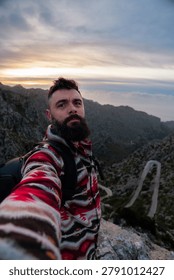 Man with a beard and backpack posing for a selfie on a rocky mountain peak overlooking a winding road and distant horizon at sunset, embodying adventure and travel