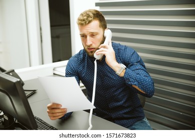 A Man With A Beard In A Blue Shirt Sits At A Computer And Talks On The Phone. He Holds A Piece Of Paper In His Hands