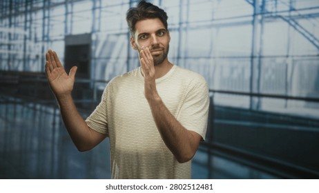 Man with beard clapping hands and smiling in airport concourse beside glass terminal windows and railing; quiet optimism.