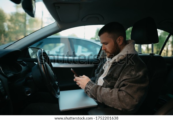 A man with a beard doing business online on his\
smartphone inside a comfort car, a laptop computer lies on his lap.\
A guy stopped his car to immediately remotely solve tasks at work\
in social distance