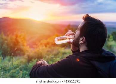 Man with beard drinking water from plastic bottle and enjoying mountain sunset. He is waring black backpack and black sport sweater. - Powered by Shutterstock