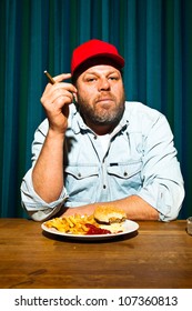 Man With Beard Eating Fast Food Meal. Enjoying French Fries And A Hamburger. Smoking A Cigar. Trucker With Red Cap.