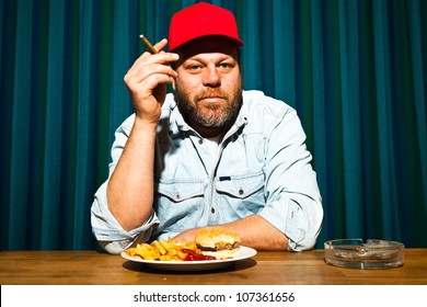 Man With Beard Eating Fast Food Meal. Enjoying French Fries And A Hamburger. Smoking A Cigar. Trucker With Red Cap.