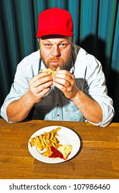 Man With Beard Eating Fast Food Meal. Enjoying French Fries And A Hamburger. Trucker With Red Cap.