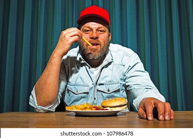 Man With Beard Eating Fast Food Meal. Enjoying French Fries And A Hamburger. Trucker With Red Cap.