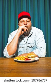 Man With Beard Eating Fast Food Meal. Enjoying French Fries And A Hamburger. Smoking A Cigar. Trucker With Red Cap.