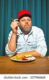 Man With Beard Eating Fast Food Meal. Enjoying French Fries And A Hamburger. Smoking A Cigar. Trucker With Red Cap.