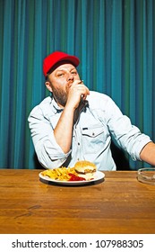 Man With Beard Eating Fast Food Meal. Enjoying French Fries And A Hamburger. Smoking A Cigar. Trucker With Red Cap.