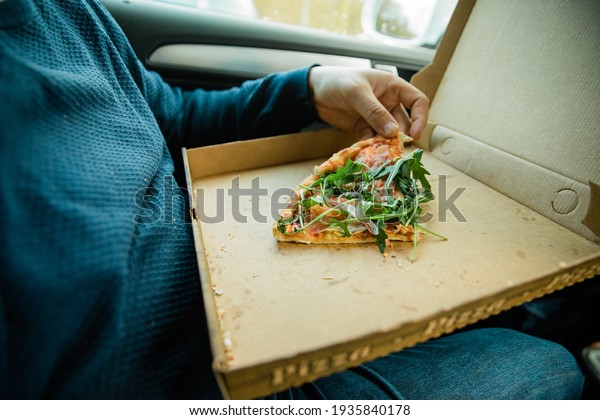 Man with beard eats pizza in his cart. Eat takeaway food\
in the car. Due to closed restaurants during the pandemic, you are\
forced to order food. Flatbread in a cardboard box that says\
\