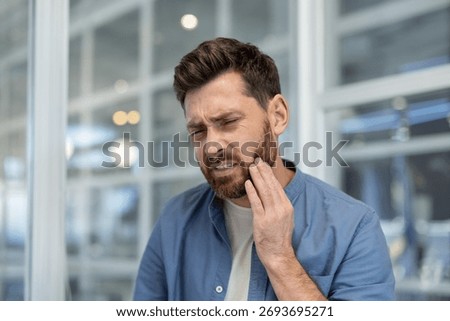 Man with beard experiencing sharp tooth pain, holding cheek with hand, frowning, expressing discomfort from a dental problem or oral health discomfort