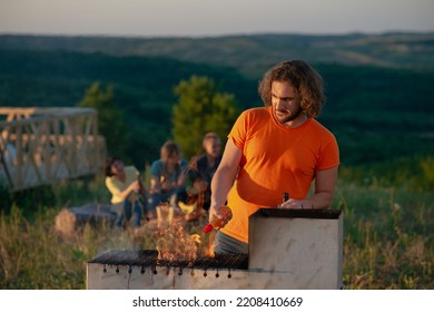 A Man With A Beard Is Getting The Fire Going At An Outside Grill While His Friends Sitting Behind Him Are Having A Good Time Out In The Fields