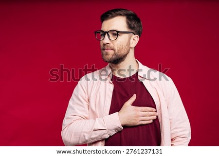 Man with beard and glasses holding hand on chest, expressing sincerity, honesty, allegiance, and patriotism on a red background