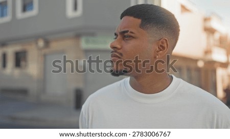 Man with beard and goatee looking left in street wearing white tshirt and earring, short fade haircut near storefront building; quiet contemplation.