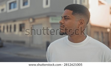 Man with beard and goatee looking left in street wearing white tshirt and earring, short fade haircut near storefront building; quiet contemplation.