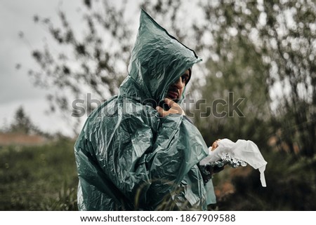 A man with a beard in a green raincoat eats in nature. Cloudy weather, fast food