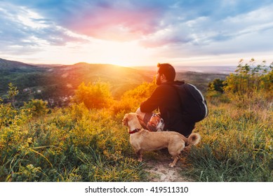 Man with beard and his small yellow dog enjoying mountain sunset and looking at the distance. He is waring black backpack and black sport sweater.