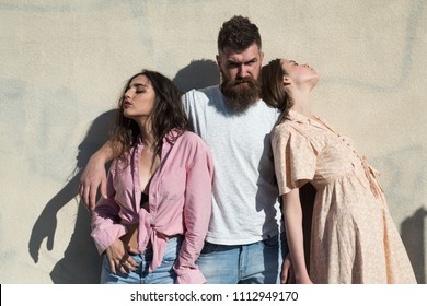 Man with beard hugs two ladies on hot sunny day. Girls turned to opposite sides while man hugs them. Love triangle concept. Threesome suffers of heat on summer day, wall on background.