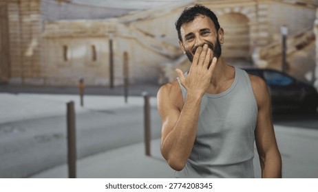 Man with beard laughing while hand covers mouth on a sunlit street by a stone building; joy carefree lightheartedness.