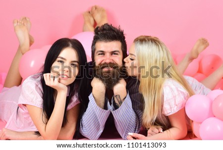 Man with beard and mustache attracts blonde and brunette girls. Threesome on smiling faces lay near balloons. Girls fall in love with bearded macho, pink background. Alpha male concept.