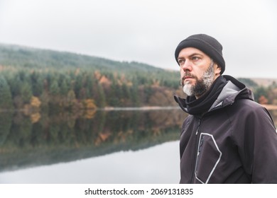 Man with beard portrait outdoor in autumn - Adult man wearing warm clothes and wool hat standing by a lake and looking away - Autumn style portrait