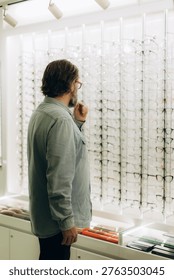Man with a beard standing in front of a display rack, carefully selecting new eyewear frames at an optical store
