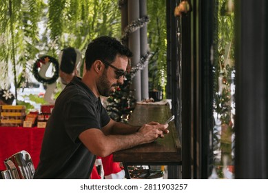 Man with beard and sunglasses using a mobile phone at a wooden counter in a cafe decorated for Christmas