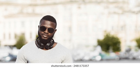 A man with a beard and sunglasses is wearing headphones around his neck. He is looking directly at the camera while standing in front of a blurred city background. - Powered by Shutterstock