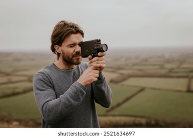 A man with a beard using a vintage camera outdoors. The man is focused on filming with the vintage camera. The vintage camera is held by the man in a rural setting. Rural autumn nature landscape.