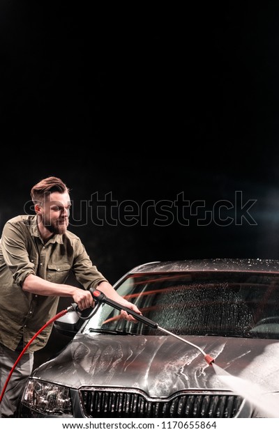 A man with a beard washes a gray car with a\
high-pressure apparatus at night in a car wash. Expensive\
advertising photography