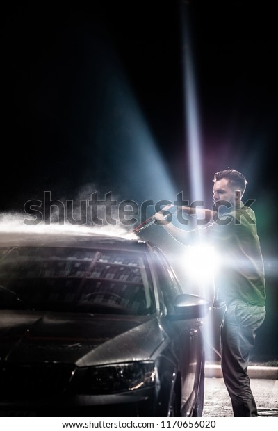 A man with a beard washes a gray car with a
high-pressure apparatus at night in a car wash. Expensive
advertising photography
